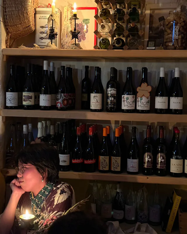 The wine wall by candlelight, shelves of small-producer bottles glowing warm while a guest rests her chin on her hand