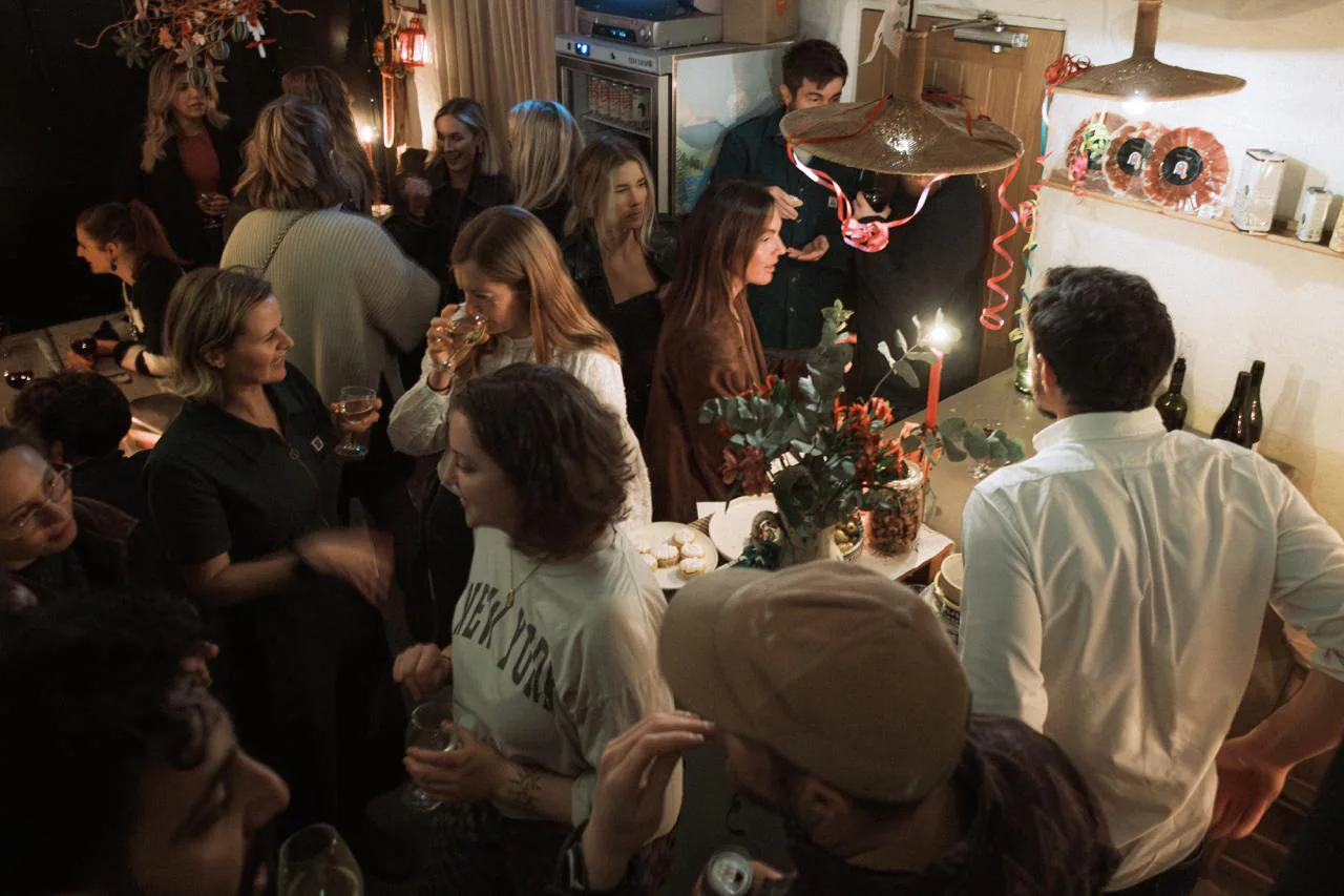 The bar full on a winter evening, friends shoulder to shoulder under warm lamplight with wine, candles and plates being passed around