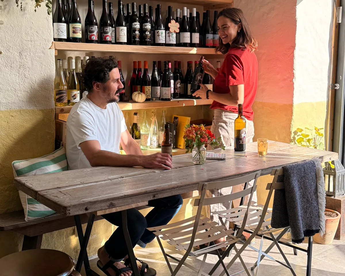 The long table in calm daylight, Ernest sitting with a bottle while Marisa sorts the wine shelves behind