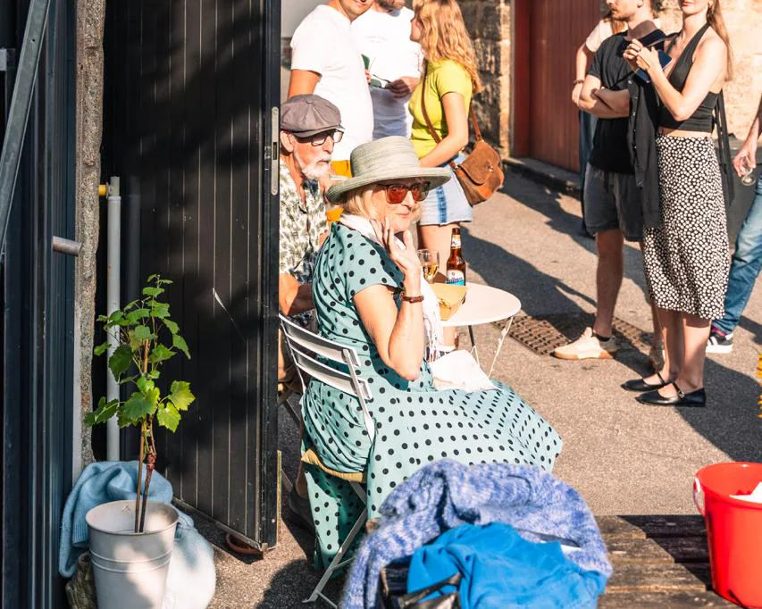A couple in sun hats sharing a drink at a little table beside the black front door, evening sun across the lane