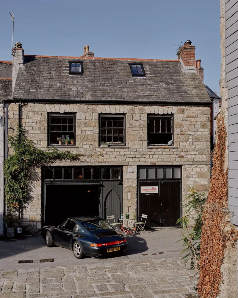 The granite frontage of The Neighbourhood on a Penryn back lane, with its red sign by the door and a classic car on the cobbles in the sun