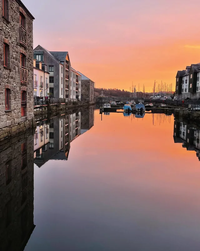 Penryn harbour at sunset, old quayside buildings and moored boats mirrored in the still water beneath a peach sky